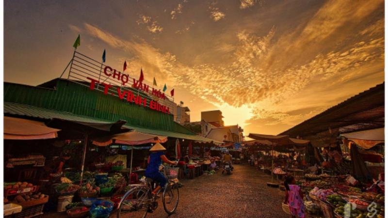 Một góc chợ dân sinh truyền thống Việt Nam nhộn nhịp, với người bán và người mua đang tương tác, xung quanh là các sạp hàng bày bán rau củ, thịt cá đầy màu sắc, thể hiện văn hóa mua sắm đặc trưng.